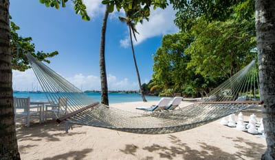 A string hammock strung between palm trees on a white-sand beach at Calabash Luxury Boutique, with sun loungers, a giant chess set, and a turquoise bay dotted with sailboats behind, Grenada.