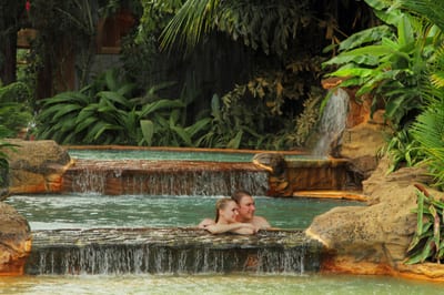 A couple soaking at the edge of a terraced hot-spring pool with water spilling over the stone lip, framed by rainforest, at the Cascadas Calientes pools of The Springs Resort in Arenal, Costa Rica