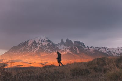 Hiker crossing golden grassland below the snow-capped granite spires of Torres del Paine at sunset, Patagonia, Chile